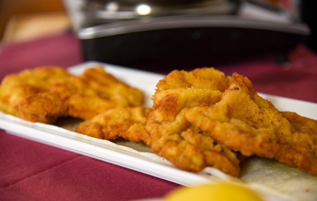 Das Bild zeigt eine Platte mit mehreren frisch zubereiteten Wiener Schnitzeln. Die Schnitzel haben eine goldene, knusprige Panade und sind gleichmäßig gebräunt. Sie sind auf einem weißen Teller angerichtet, der auf einem rot gedeckten Tisch steht. Im Vordergrund ist ein Stück Zitrone sichtbar, das anscheinend zum Anrichten oder zum Servieren der Schnitzel gehört. Der Hintergrund des Bildes ist unscharf, zeigt aber eine Art Kochutensil oder eine wiederverwendbare Küchenausstattung, die sich im oberen Bereich des Bildes befindet.