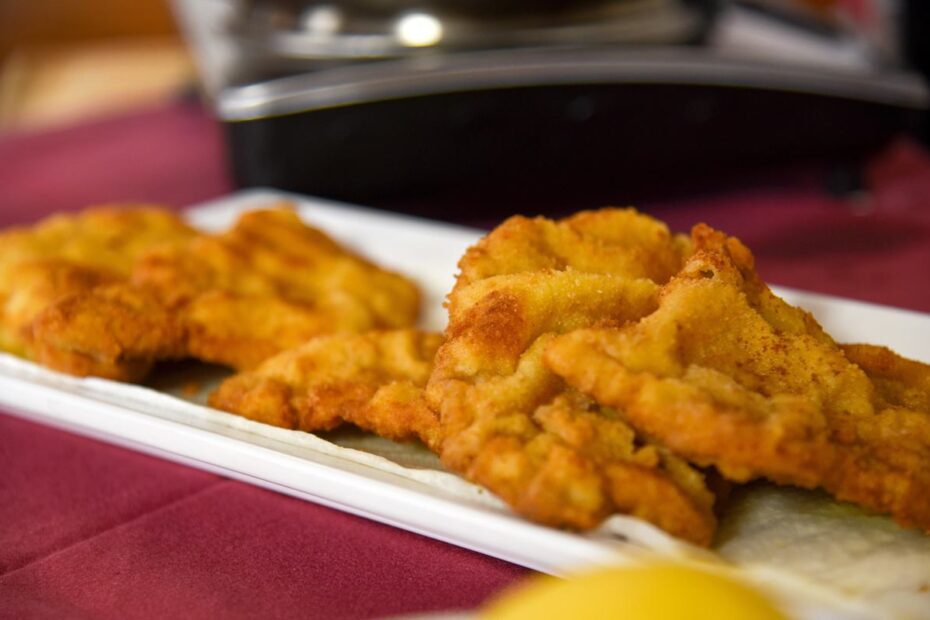 Das Bild zeigt eine Platte mit mehreren frisch zubereiteten Wiener Schnitzeln. Die Schnitzel haben eine goldene, knusprige Panade und sind gleichmäßig gebräunt. Sie sind auf einem weißen Teller angerichtet, der auf einem rot gedeckten Tisch steht. Im Vordergrund ist ein Stück Zitrone sichtbar, das anscheinend zum Anrichten oder zum Servieren der Schnitzel gehört. Der Hintergrund des Bildes ist unscharf, zeigt aber eine Art Kochutensil oder eine wiederverwendbare Küchenausstattung, die sich im oberen Bereich des Bildes befindet.