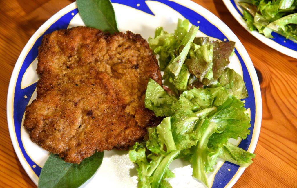 Das Bild zeigt einen Teller mit einer Cotoletta alla Milanese, einem italienischen Schnitzel. Das Schnitzel ist goldbraun und knusprig, auf einem blauen Teller mit einem einfachen, aber dekorativen Rand arrangiert. Neben dem Schnitzel liegt ein frischer Salat mit grünen Blättern, der ebenfalls auf dem Teller serviert ist. Im Hintergrund sind weitere Teller mit Salat und anderen Speisen sichtbar. Der Tisch, auf dem die Teller stehen, ist aus Holz und hat eine warme, natürliche Farbe.