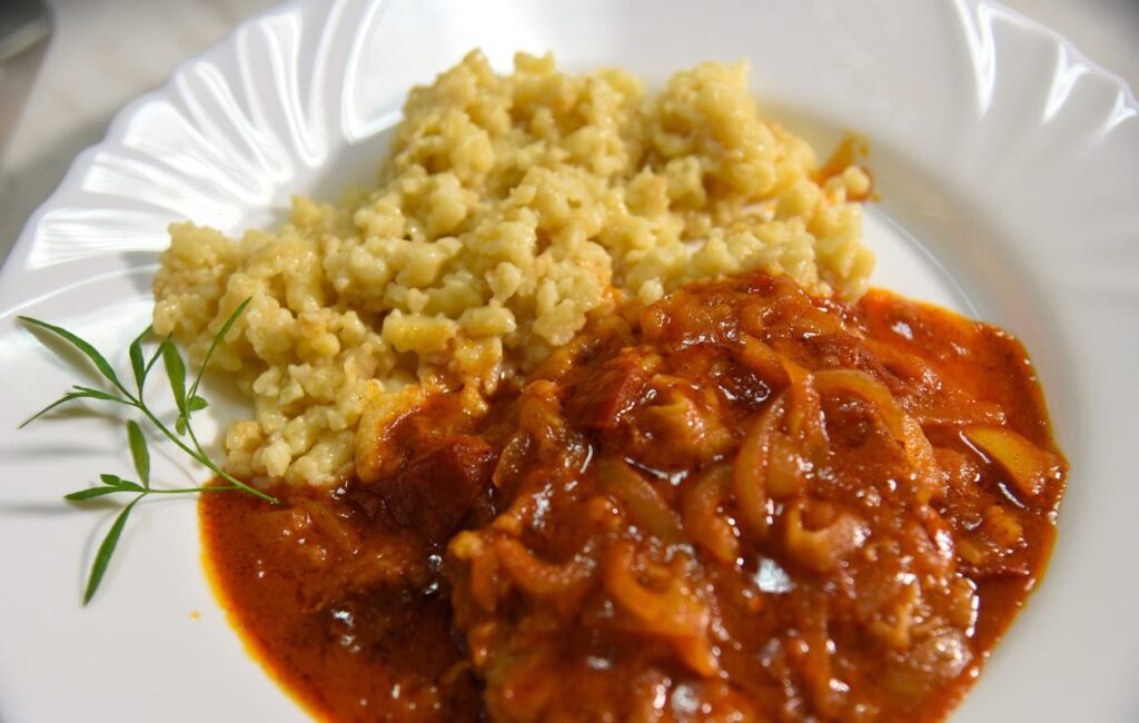 Das Bild zeigt einen Teller mit einem Gericht, das aus zwei Hauptbestandteilen besteht. Links sind Stücke von Fleisch zu sehen, die in einer dicken, roten Sauce serviert werden. Die Sauce hat eine glatte Konsistenz und eine würzige Farbe, und es sind einige fleischliche Zutaten sichtbar. Rechts vom Fleisch befindet sich eine Portion von Tarhonya, kleinen, genussfertigen Teigwaren, die hellgelb bis beige gefärbt sind. Diese sind weich und haben eine unregelmäßige Form. Der Teller, auf dem die Speisen angerichtet sind, hat einen klassischen, weißen Rand, der das Essen hervorhebt. Das gesamte Gericht wirkt frisch zubereitet und scheint sehr reichhaltig und schmackhaft zu sein. Hintergrund ist unscharf, sodass der Fokus klar auf dem Essen liegt.
